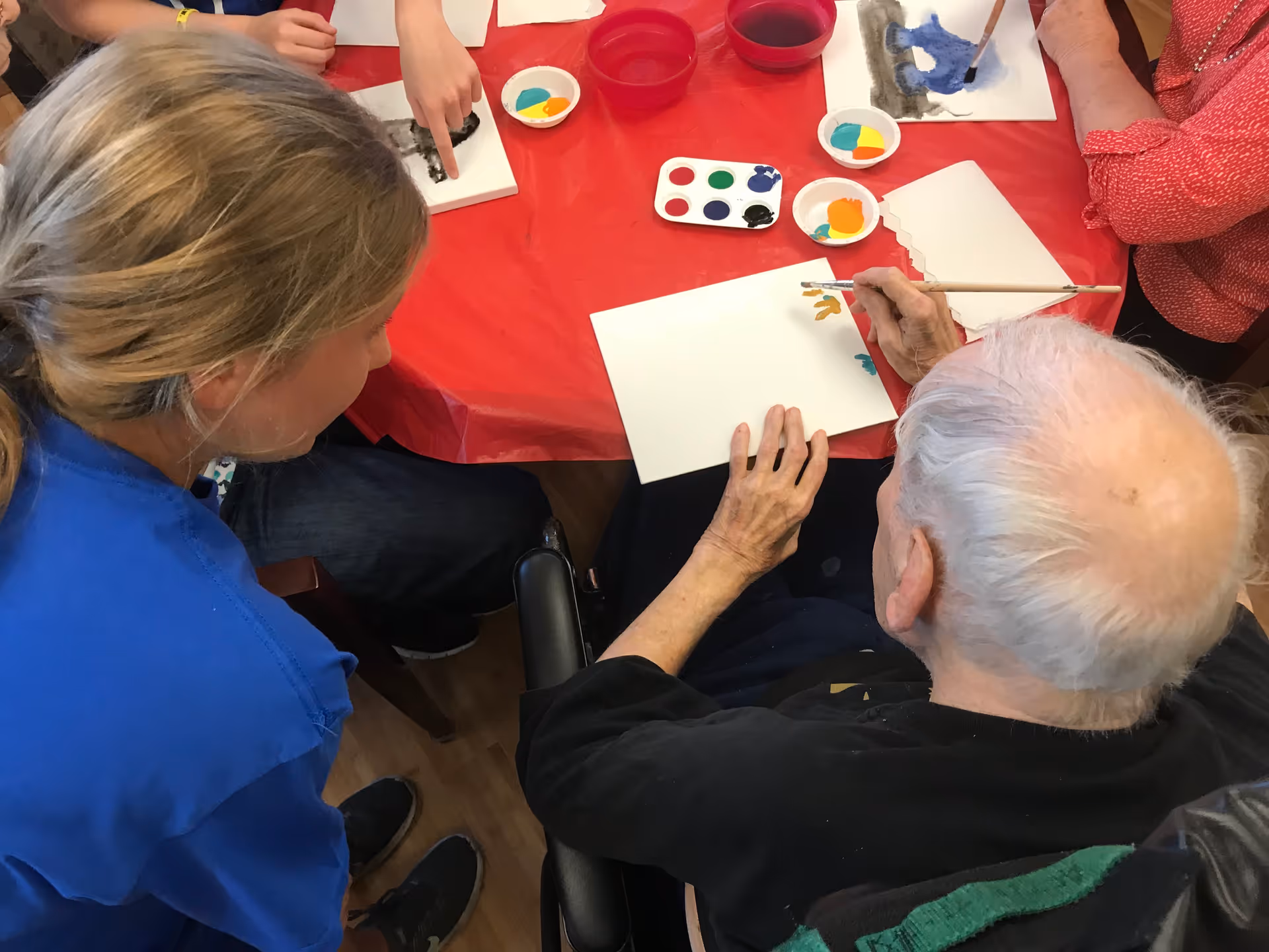 An elderly man seated at a table is painting on a white canvas with a brush, while a woman in a blue shirt leans in to assist or observe. The table is covered with a red tablecloth and has paint palettes and cups of water on it. Other people are partially visible around the table also engaged in painting activities.