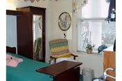 Small nursing-home bedroom with a bed, wooden dresser and wardrobe, a striped rocking chair by a window with blinds and potted plants.