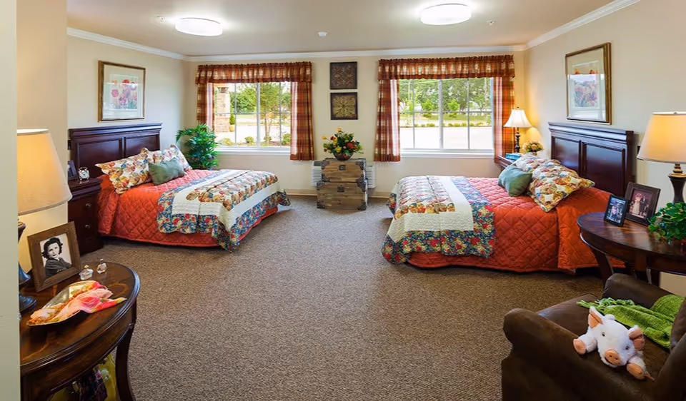 Bright shared bedroom with two twin beds, bedside tables, lamps, and large windows overlooking greenery.