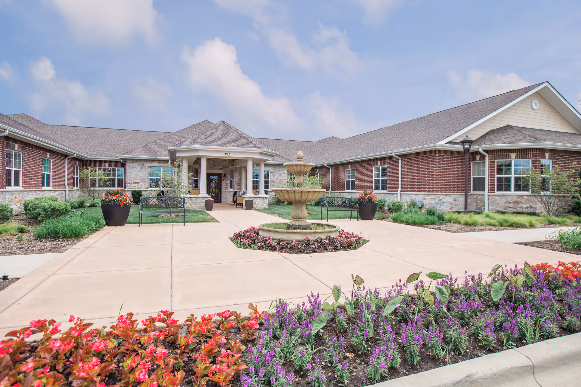Exterior view of a single-story brick and stone building with a covered entrance, a central fountain, benches, and landscaped flower beds with colorful flowers under a partly cloudy sky.