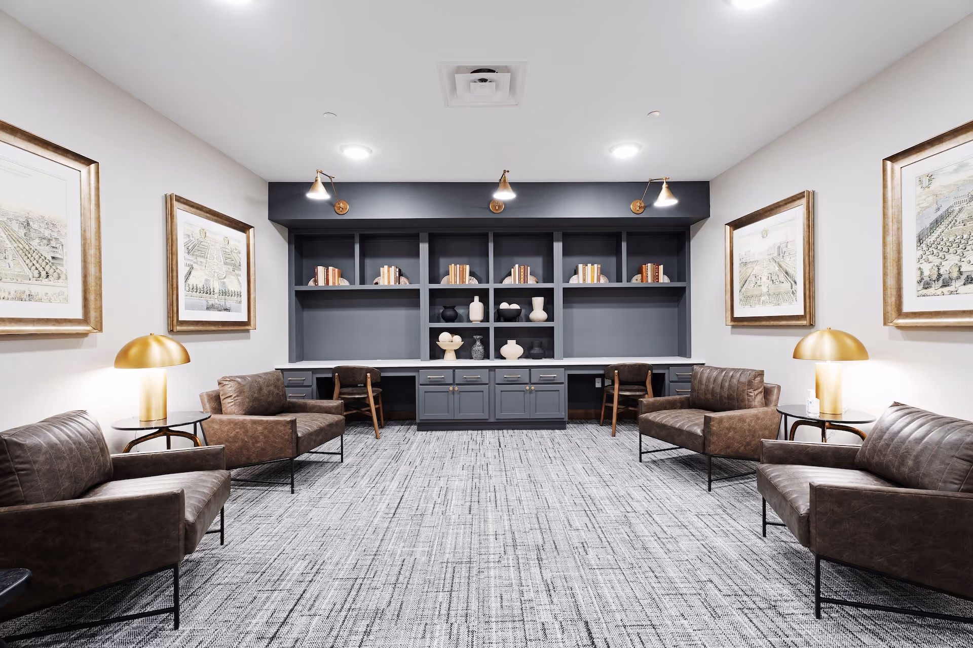 A modern lounge area with four brown leather armchairs arranged around two small round tables with gold lamps. The back wall features a built-in dark gray shelving unit with books and decorative vases. The room has framed artwork on the walls and a patterned gray carpet.