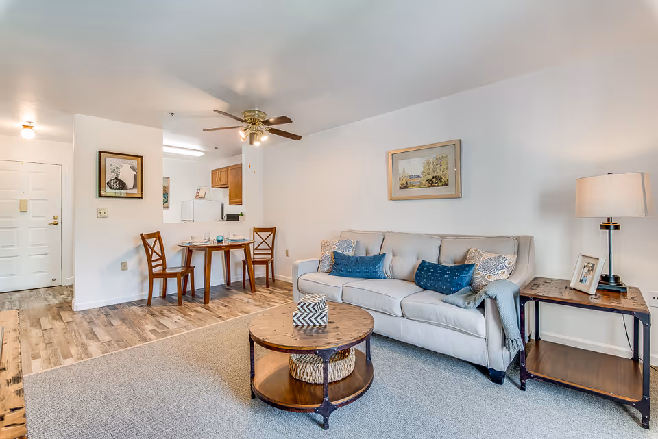 Open living room and dining area with a beige sofa, round wooden coffee table, small dining table and a visible kitchenette.