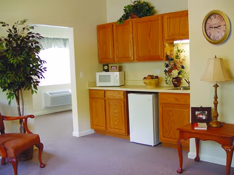 Interior view of a senior living facility room featuring a small kitchenette with wooden cabinets, a microwave, a mini refrigerator, and a countertop with a basket of fruit and a vase of flowers. To the right, there is a wooden side table with a lamp and a framed photo, and a wall clock above it. On the left side, there is a potted plant and an open doorway leading to a bright room with a window and an air conditioning unit.