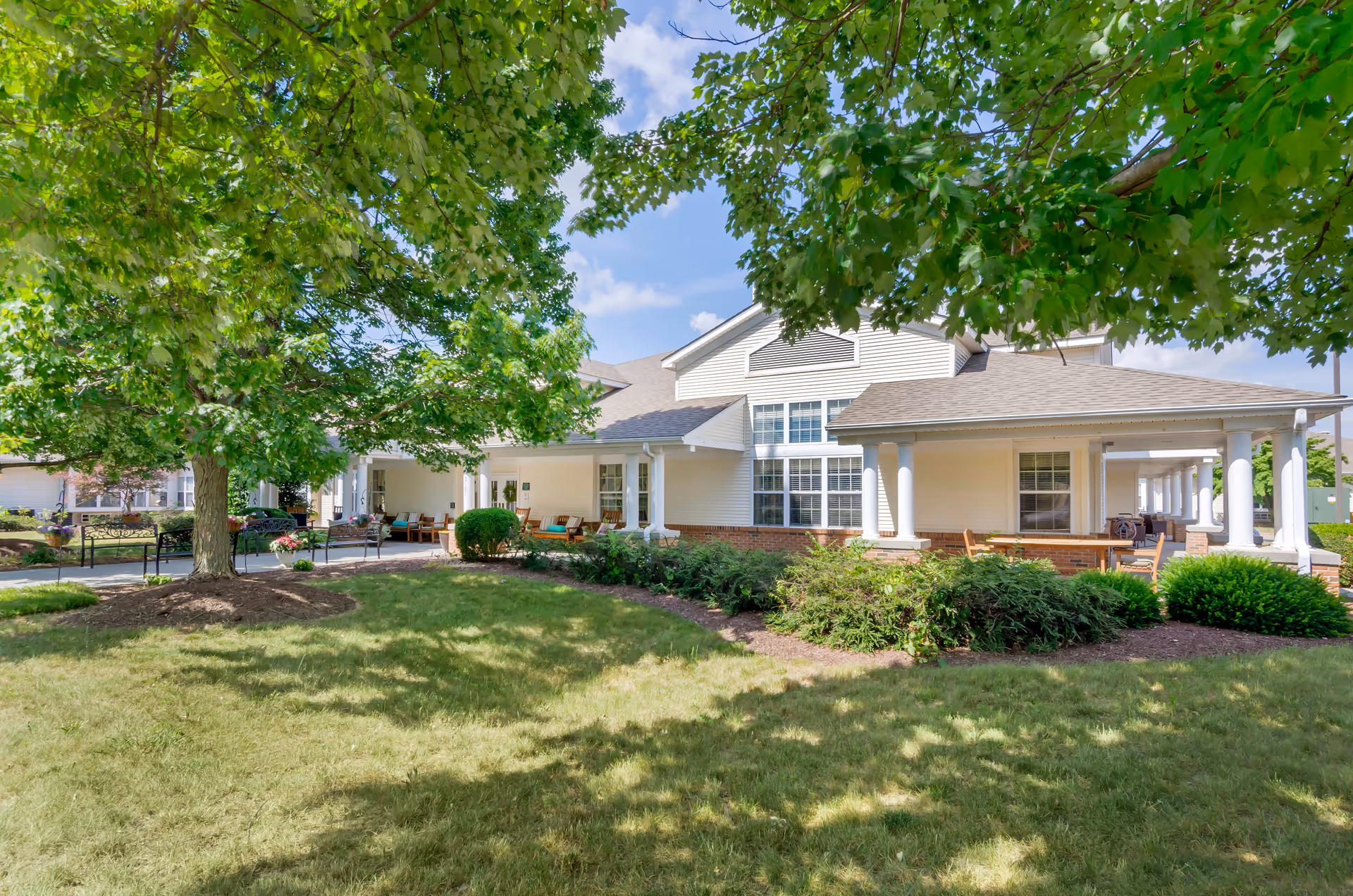 Exterior view of Brookdale Harrisonburg senior living facility showing a white building with large windows, a covered porch with seating, surrounded by green trees and shrubs under a partly cloudy sky.