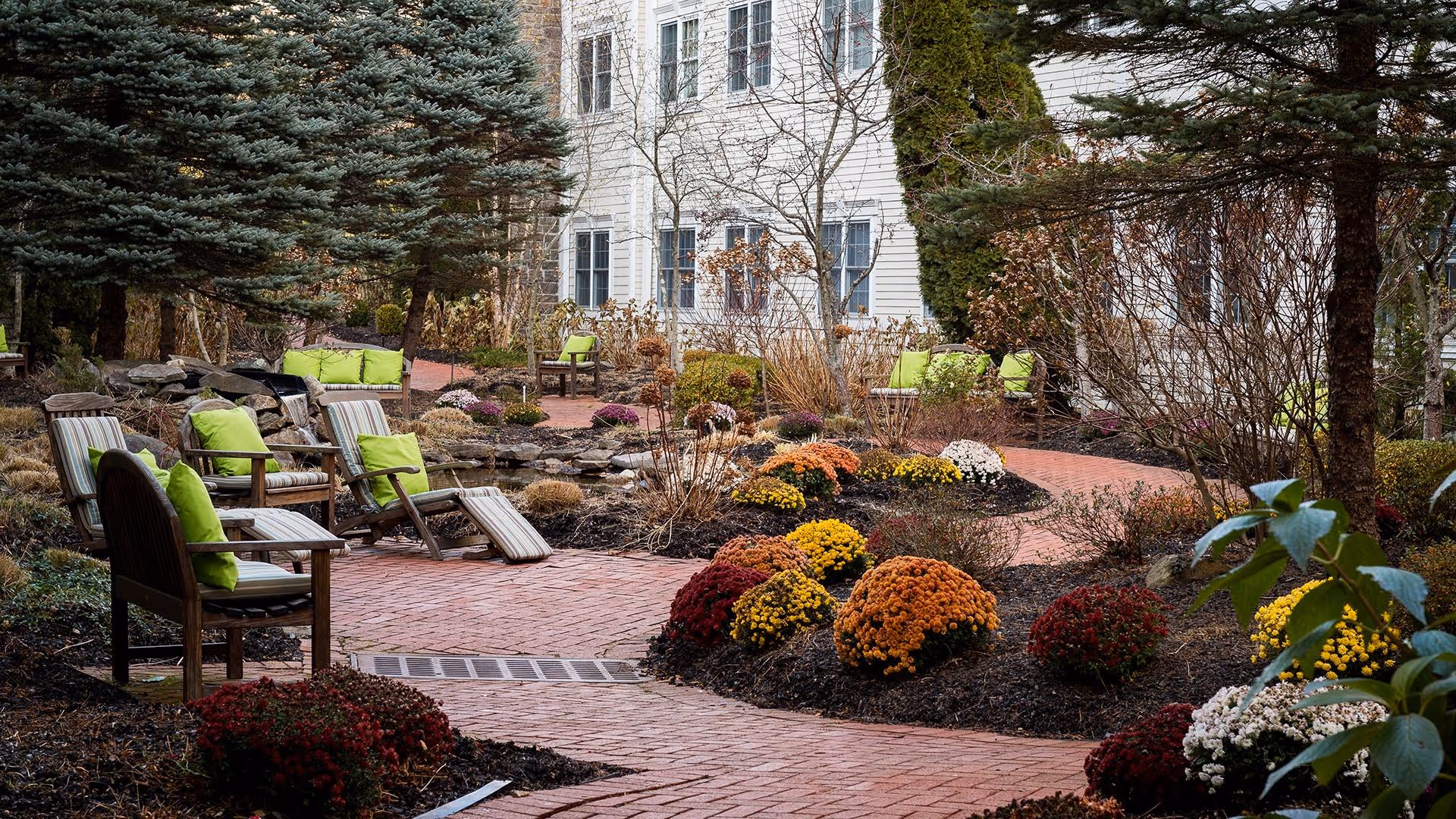 A landscaped outdoor garden area with a winding brick pathway surrounded by colorful flower bushes and trees. Several wooden chairs and lounge chairs with green cushions are placed along the path. A white multi-story building is visible in the background.