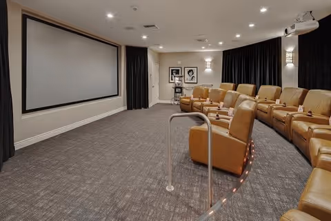 A small private movie theater room with a large blank projection screen on the front wall, rows of tan leather recliner chairs with cup holders, dark curtains on the side walls, and soft overhead lighting.