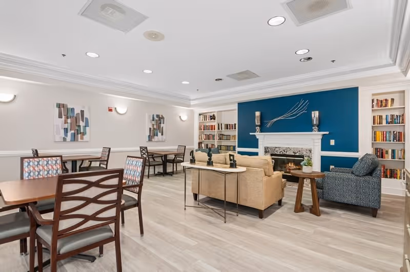 A cozy common area with a beige sofa, a blue armchair, and a white fireplace with a blue accent wall behind it. The room has bookshelves filled with books on either side of the fireplace, several tables with chairs, abstract wall art, and light wood flooring.