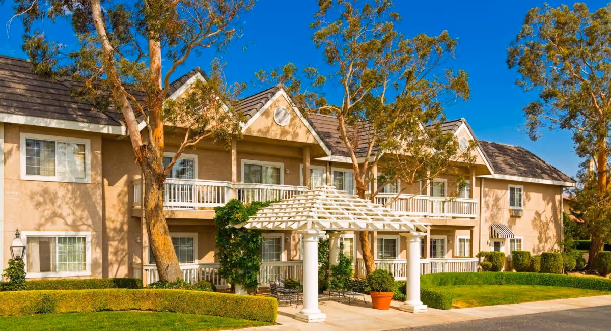 Front view of a two-story beige assisted-living building with white balconies, a pergola entrance, trees, and manicured landscaping under a blue sky.
