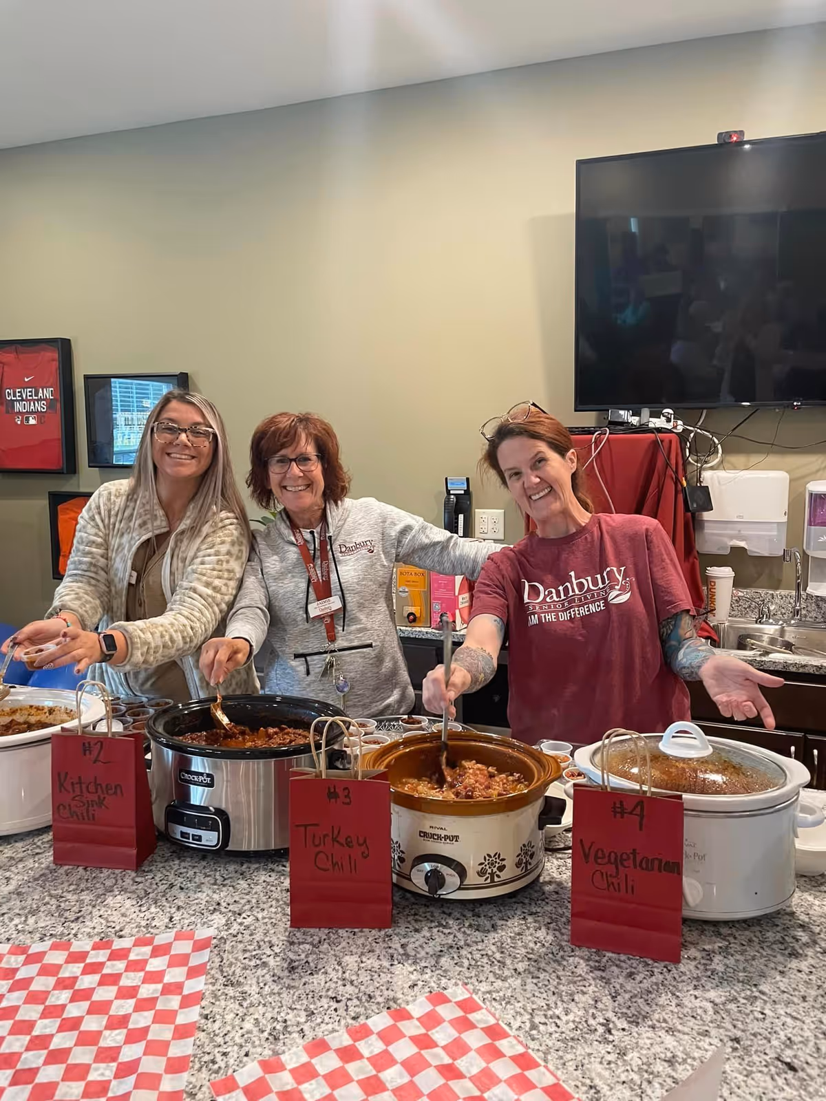 Three women standing behind a kitchen counter serving chili from crockpots labeled Kitchen Sink Chili, Turkey Chili, and Vegetarian Chili. They are smiling and appear to be in a communal or senior living facility kitchen area. One woman is wearing a maroon Danbury Senior Living t-shirt.