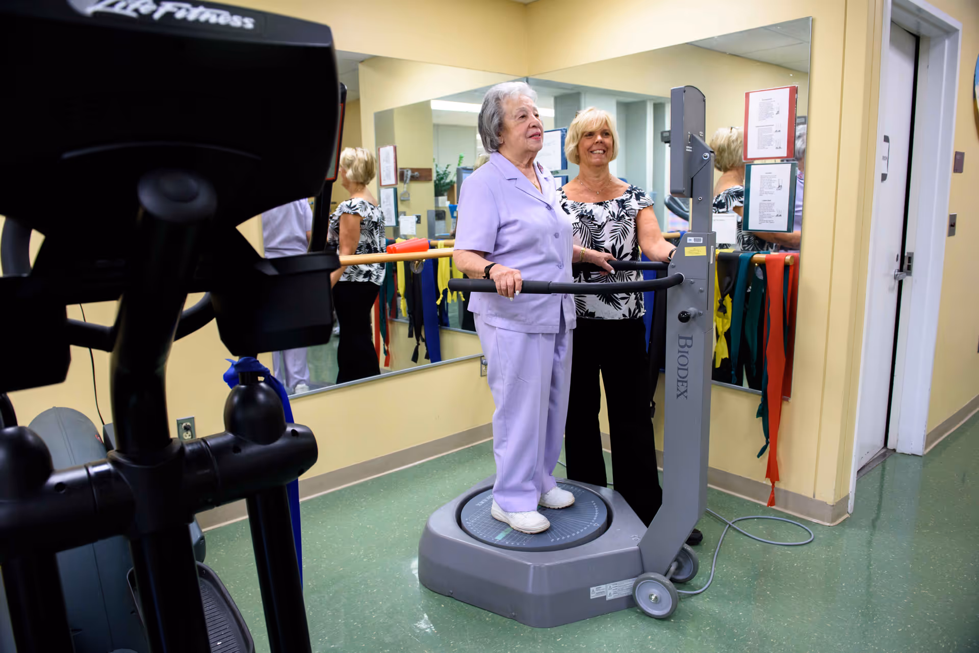 An elderly woman in a lavender outfit stands on a Biodex balance machine while a smiling woman in a black and white patterned blouse assists her in a fitness or rehabilitation room with exercise equipment and mirrors on the walls.