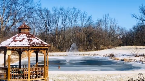 A wooden gazebo with a snow-covered roof stands near a partially frozen pond with a fountain in the center. Surrounding the pond are snow-dusted grassy areas and leafless trees under a clear blue sky.