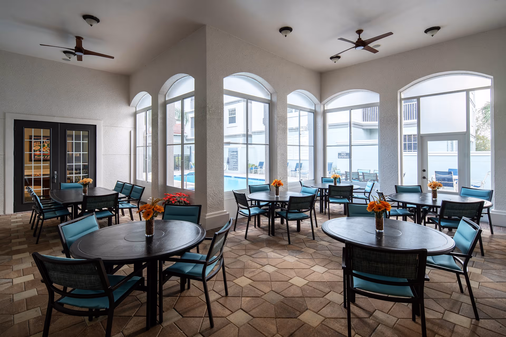 Indoor dining area with multiple round tables and teal cushioned chairs. Each table has a small vase with orange flowers. Large arched windows provide a view of an outdoor pool area. The floor has a patterned tile design, and ceiling fans are mounted above.