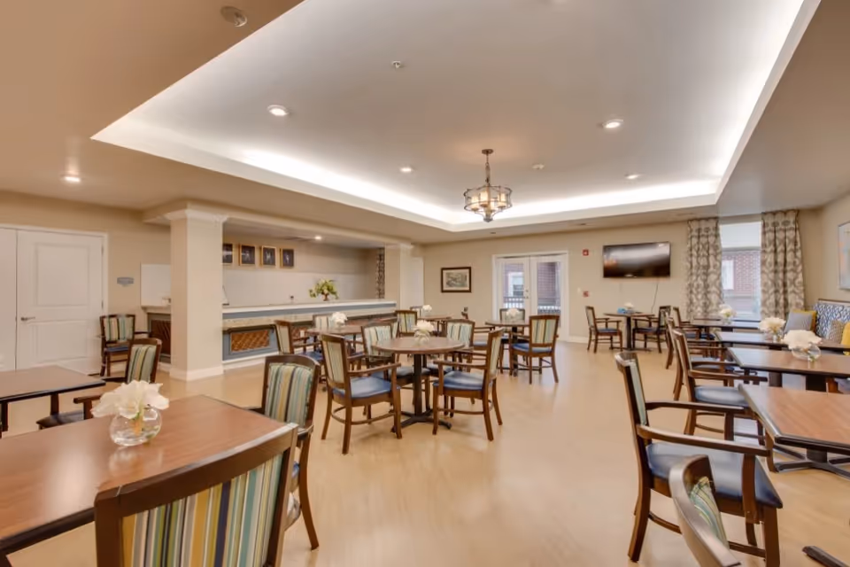 Well-lit communal dining room with multiple tables and chairs, a serving counter, and a wall-mounted TV.