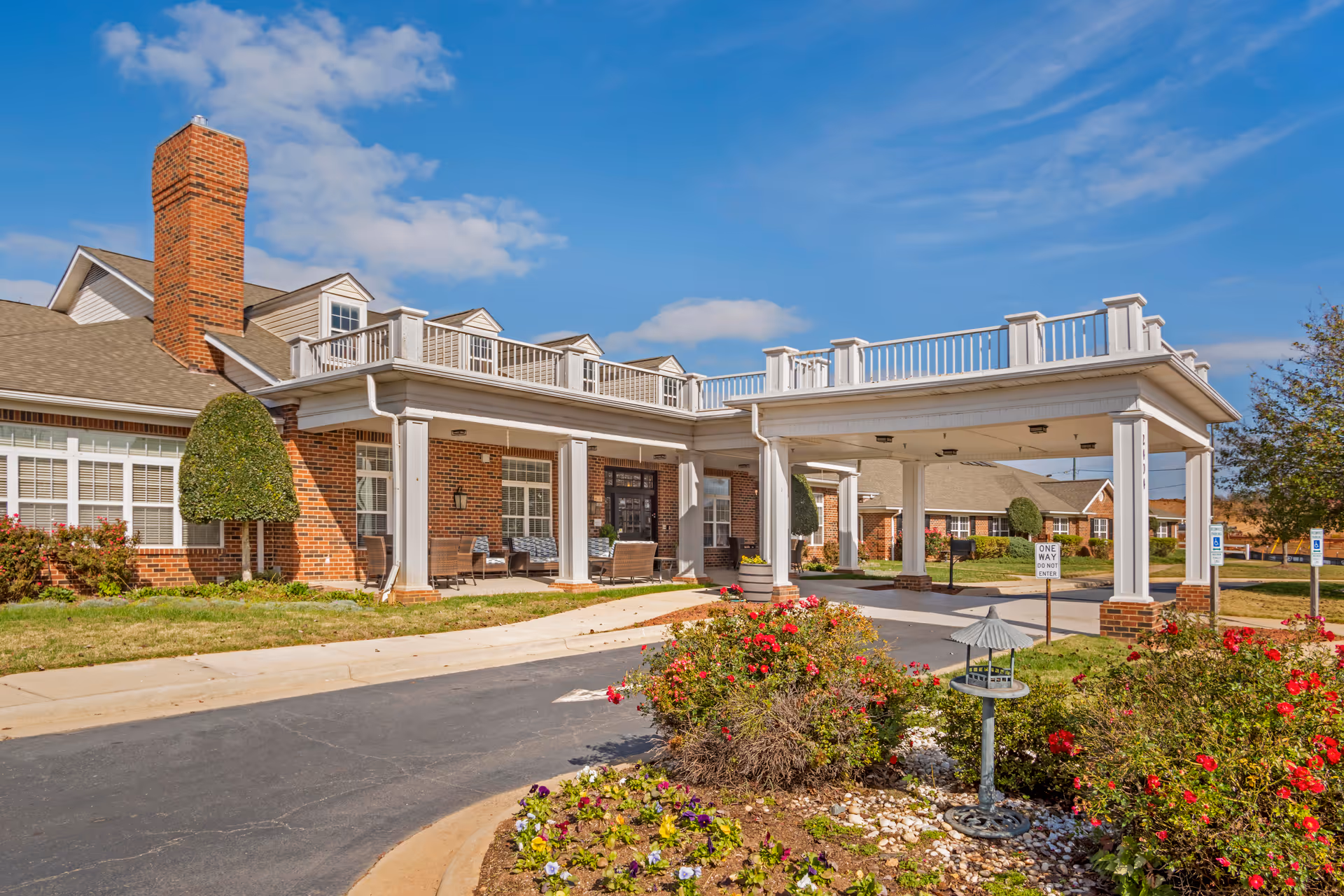 Exterior view of a senior living facility building with a covered entrance, brick walls, white columns, and a landscaped garden with flowers and shrubs in front. The sky is clear with a few clouds.