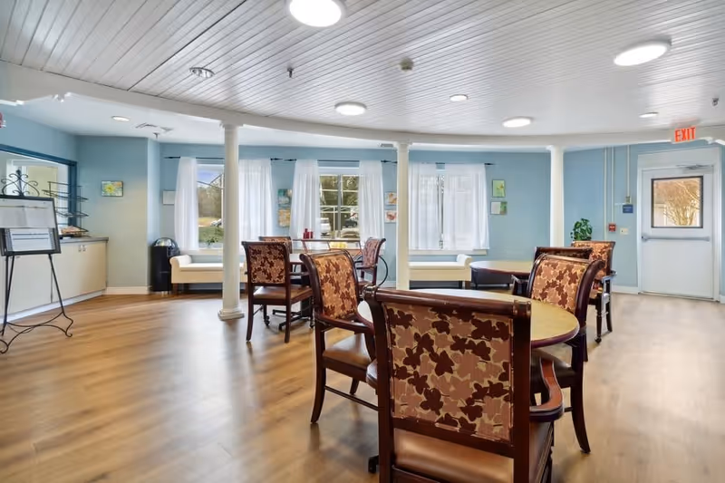 Bright communal dining room with round tables and patterned chairs, pale blue walls, columns and windows with white curtains.
