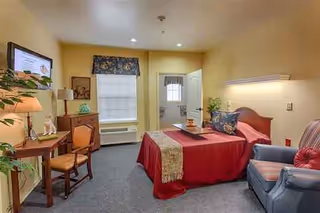 Interior of a furnished senior living room with bed, sofa, desk, and TV.