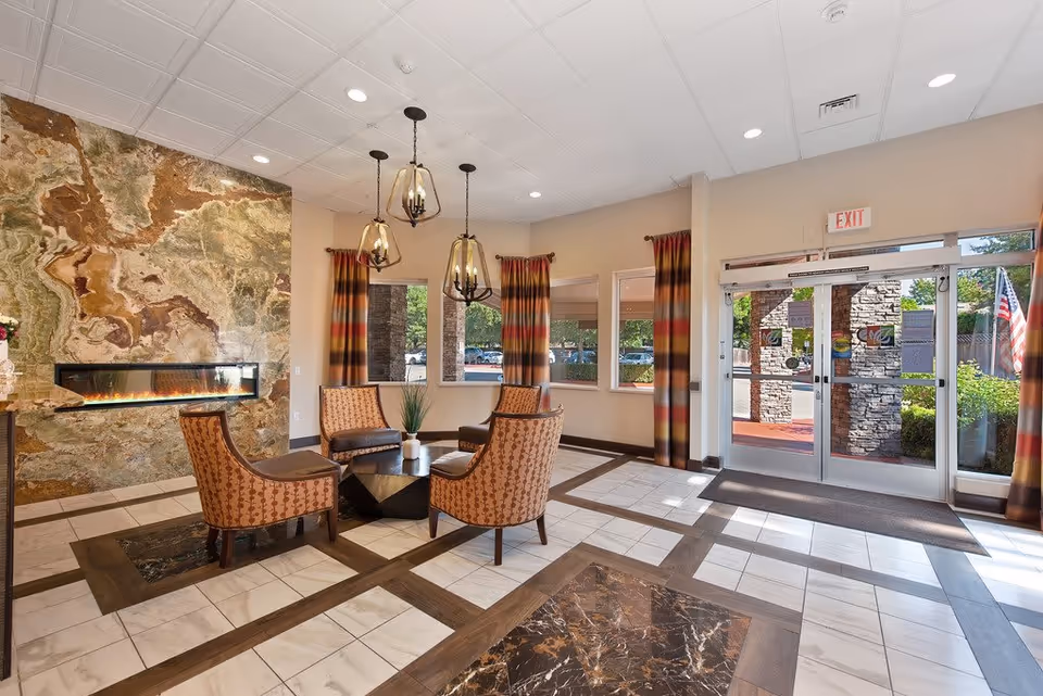 Bright senior living lobby with a seating area of patterned chairs around a coffee table, modern pendant lights, a decorative wall fireplace, and glass entrance doors.