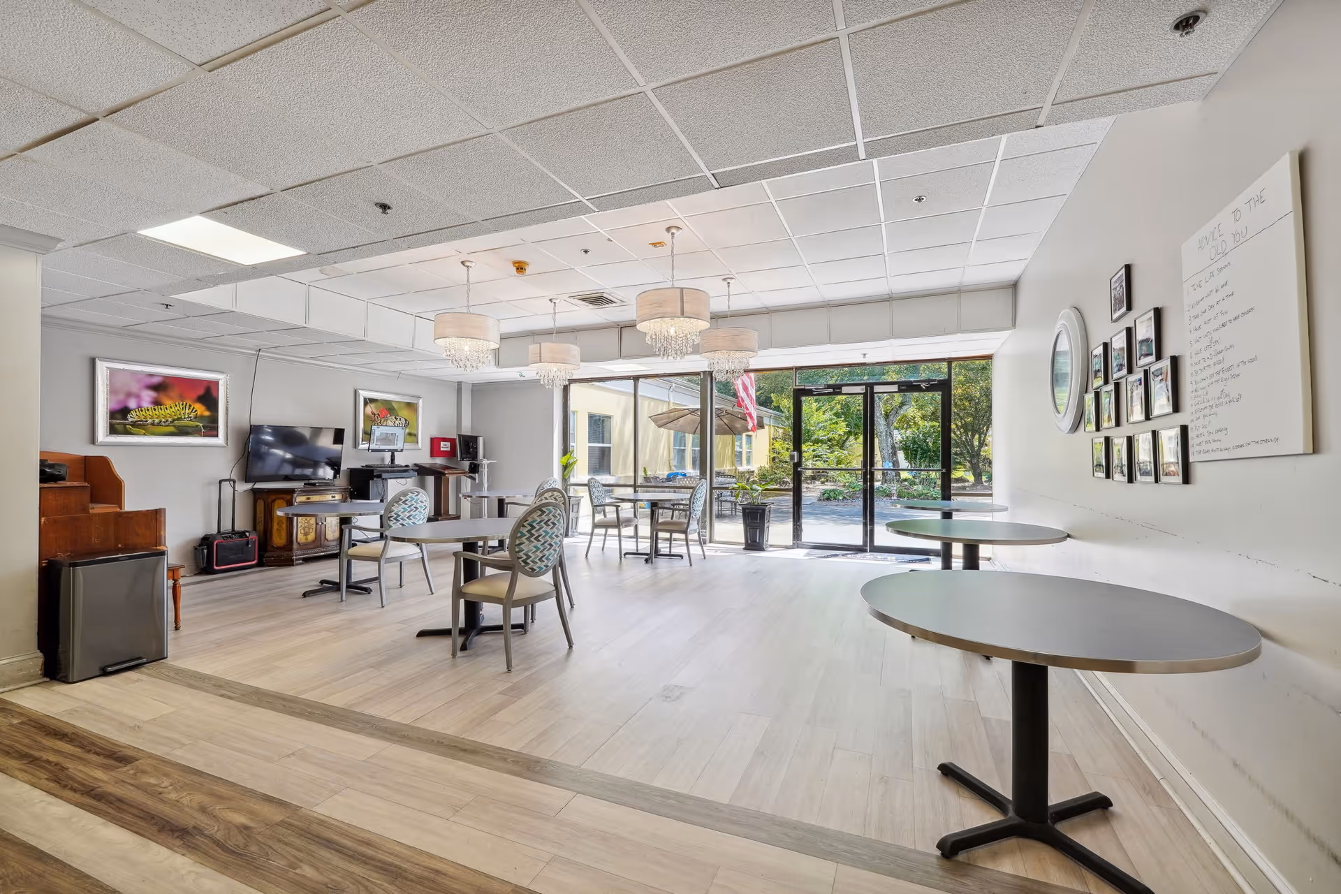 A bright and spacious common area in a senior living facility with several round tables and chairs arranged for seating. The room features large glass doors leading outside, allowing natural light to fill the space. There are three elegant chandeliers hanging from the ceiling, framed pictures on the walls, a television, and a small piano in the corner.