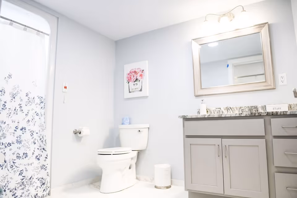 A clean and bright bathroom featuring a white toilet, a gray vanity with a marble countertop, a large framed mirror above the vanity, a floral shower curtain, and a wall decoration with pink flowers.