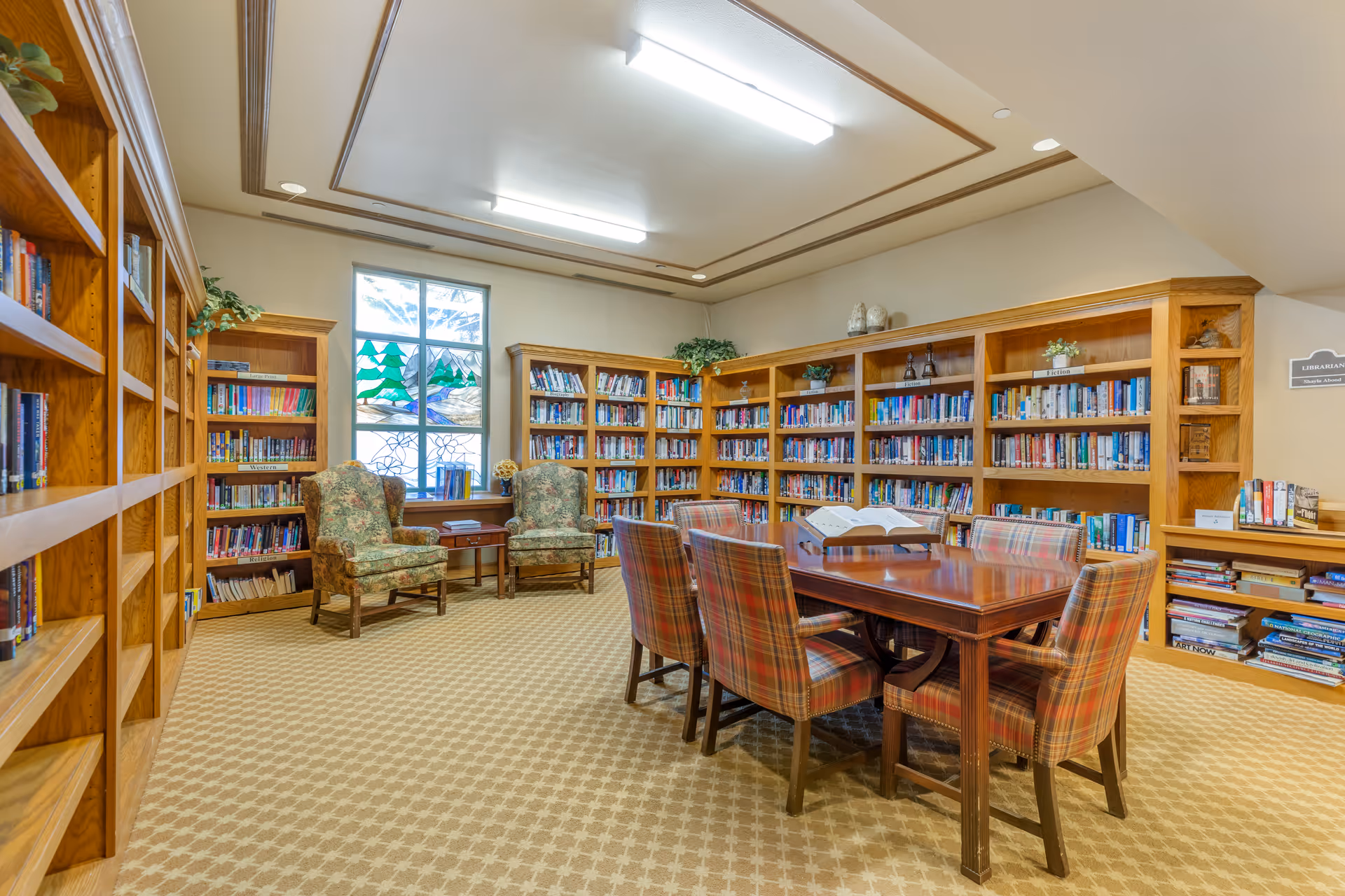 A cozy library room with wooden bookshelves filled with books lining the walls. There is a wooden table in the center surrounded by six plaid upholstered chairs. Two floral upholstered armchairs sit near a window with stained glass artwork. The room has a beige patterned carpet and soft overhead lighting.