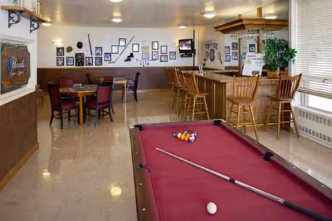 Interior view of a recreational room with a red pool table in the foreground, a wooden bar with high chairs on the right, and several tables with chairs in the background. The walls are decorated with framed pictures and memorabilia, and there is a large window with blinds on the right side.