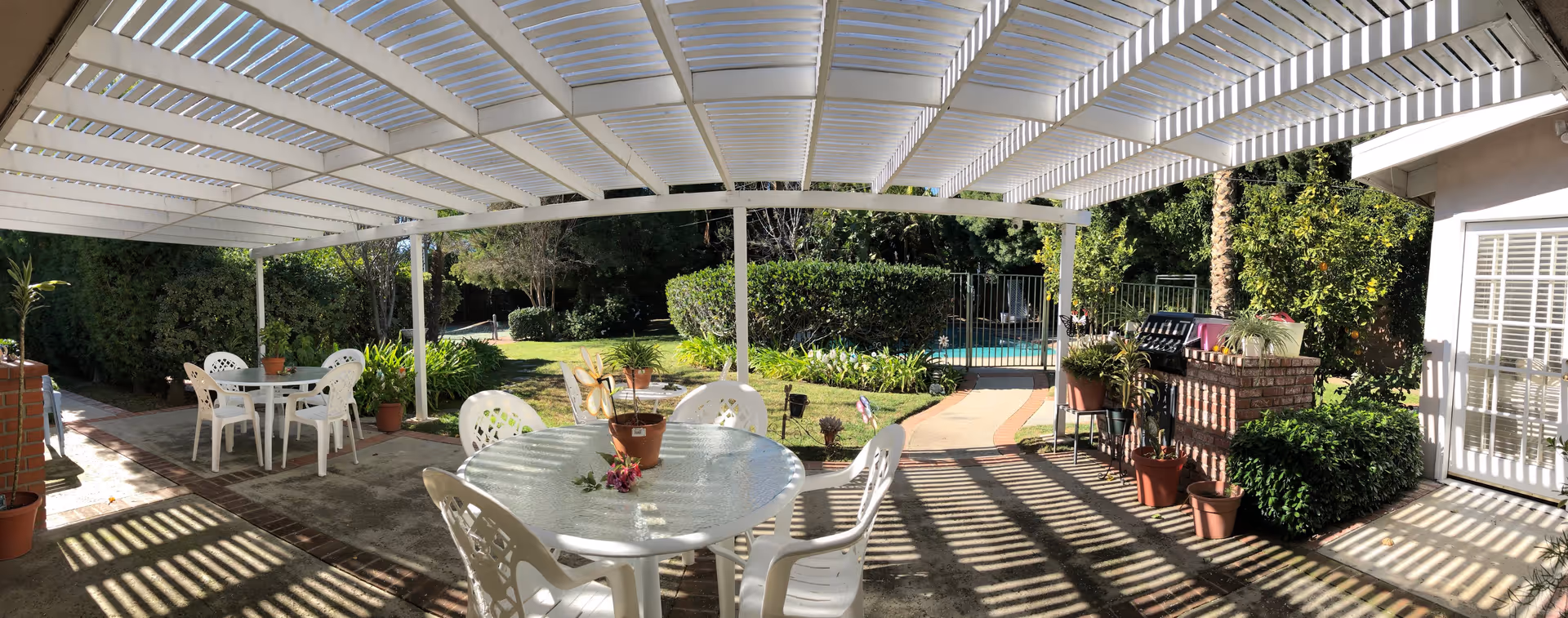 Covered outdoor patio with a white pergola, plastic tables and chairs, potted plants, and a garden with a pool visible beyond.