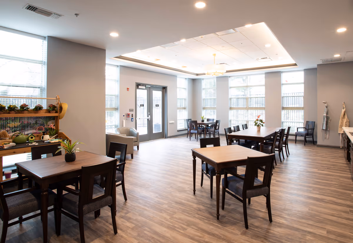 A bright and spacious dining area with multiple wooden tables and chairs arranged neatly. Large windows with blinds allow natural light to fill the room. There is a door leading outside, a small seating area with two armchairs, and a wooden shelf with potted plants and decorative items. The ceiling has recessed lighting and a central chandelier.