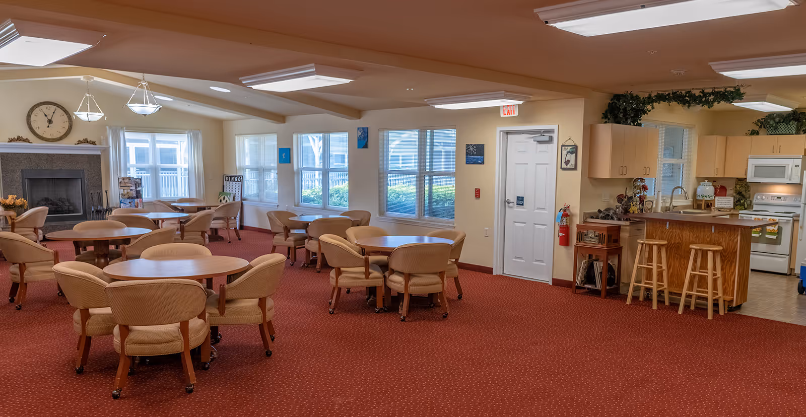 A spacious common area in a senior living facility featuring multiple round tables with cushioned chairs on a red carpet. The room has large windows allowing natural light, a fireplace with a clock above it, and a kitchen area with wooden cabinets, a microwave, stove, and a breakfast bar with stools. The walls are decorated with small paintings and greenery above the kitchen cabinets.