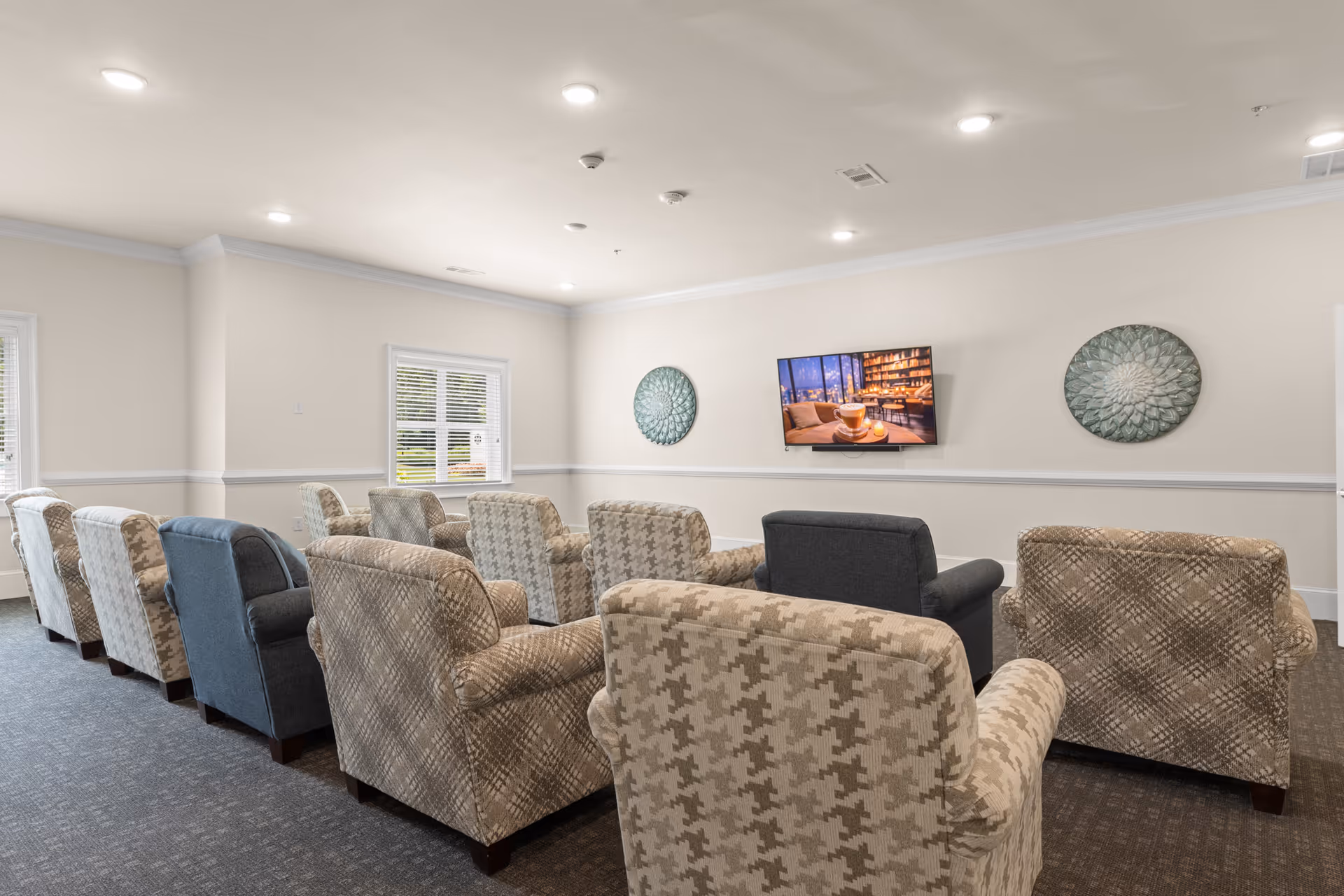 A cozy common area with multiple upholstered armchairs arranged in rows facing a wall-mounted flat-screen TV. The room has beige walls with white trim, two decorative circular wall hangings, and windows letting in natural light.