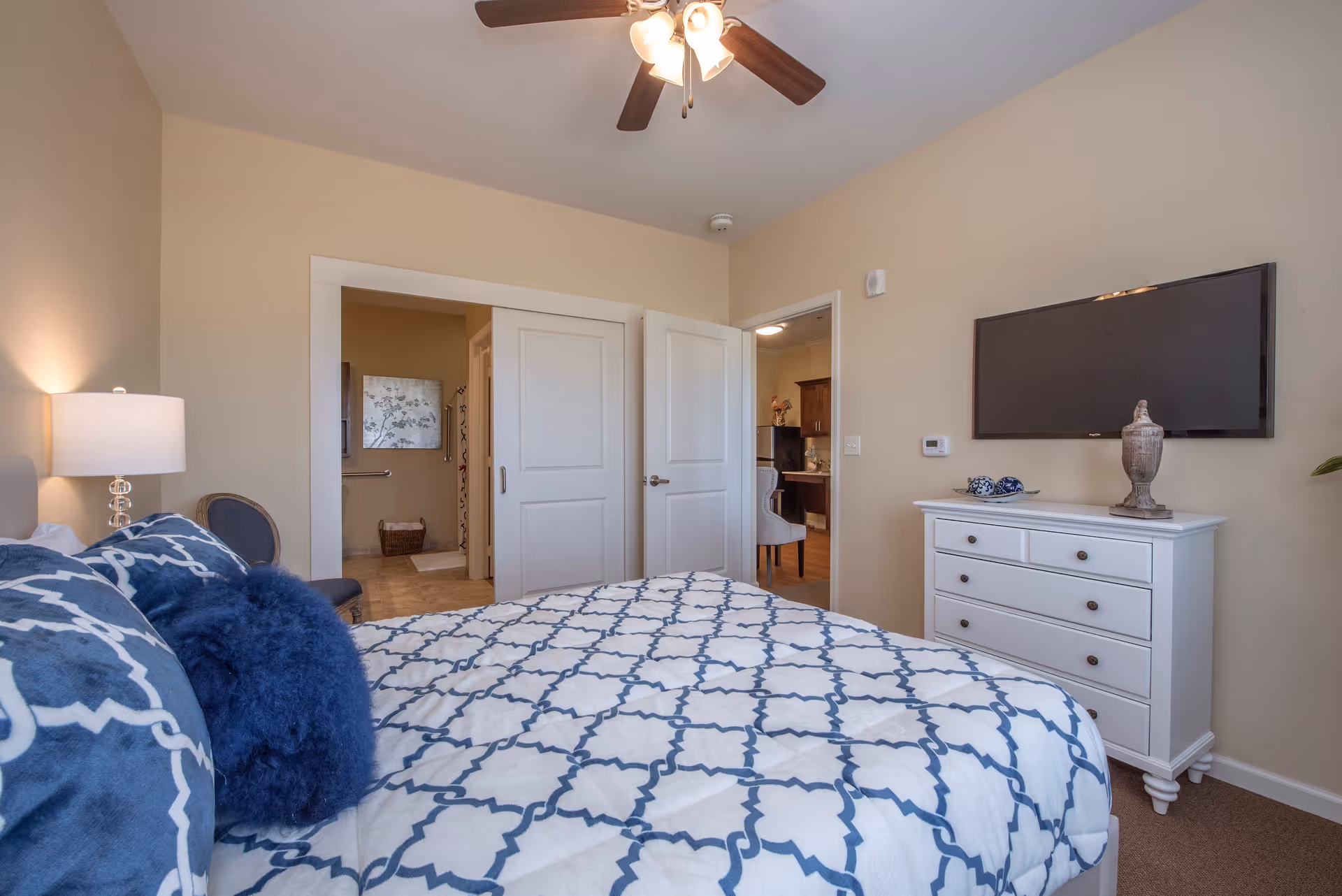 A bedroom with a bed covered in a white and blue patterned comforter and several pillows. There is a white dresser with decorative items on top and a flat-screen TV mounted on the wall above it. A ceiling fan with lights is visible on the ceiling. The room has beige walls and carpeted floor. Two doors are open, showing a glimpse of a bathroom and another room with wooden flooring and furniture.