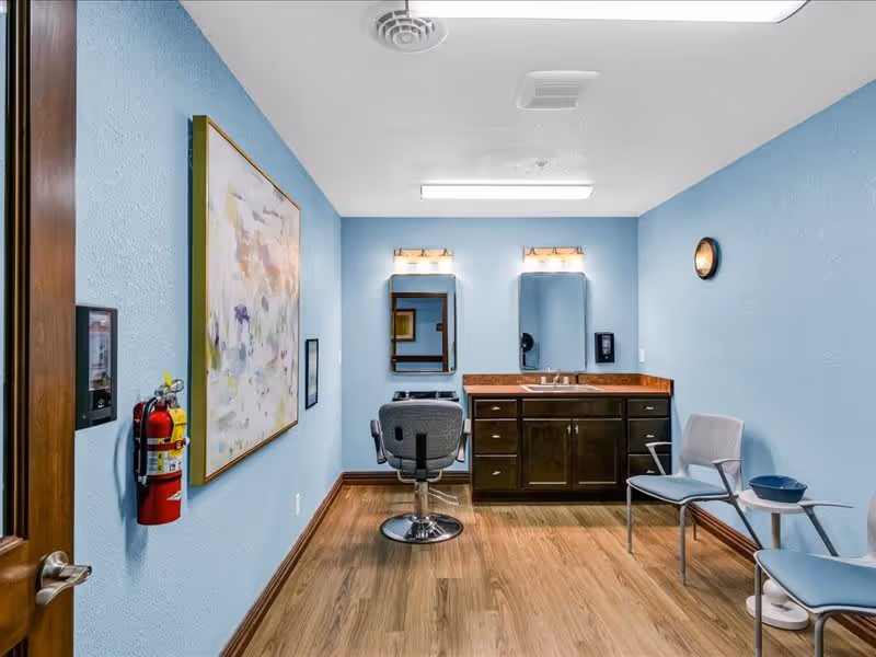 A small room with light blue walls and wood flooring featuring a salon-style chair in front of a vanity with two mirrors and two light fixtures above. There are two chairs with a small table and a bowl on the right side, a fire extinguisher mounted on the left wall, and a large abstract painting hanging on the left wall.
