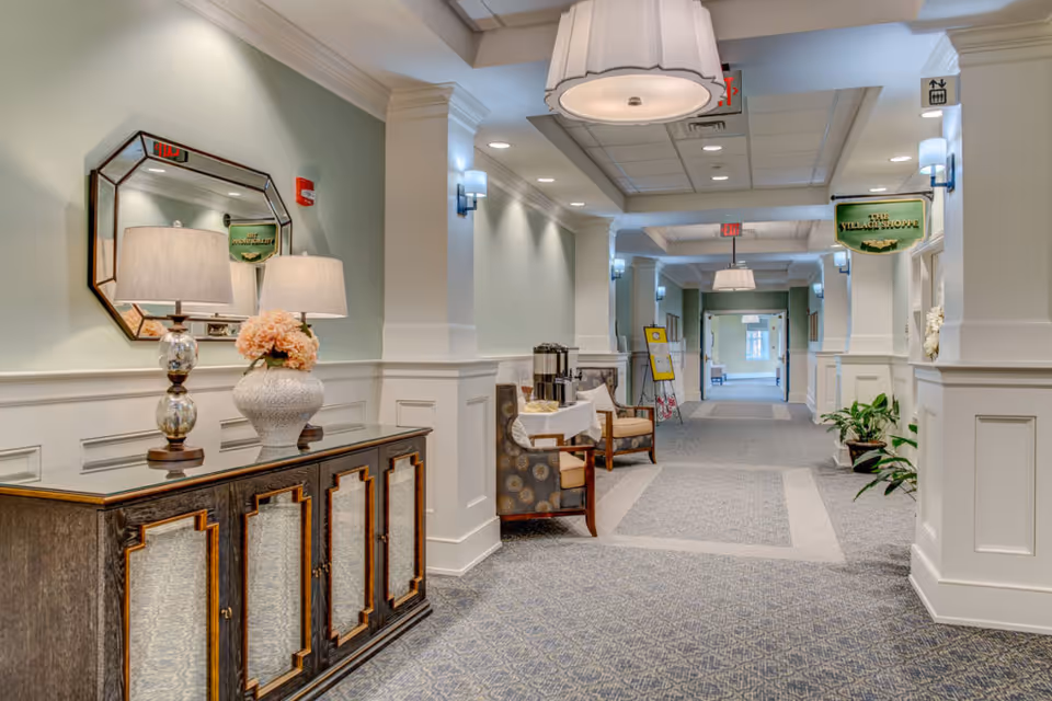 A well-lit hallway in a senior living facility with light green walls and white wainscoting. On the left, there is a dark wooden cabinet with mirrored panels, topped with two table lamps and a vase of flowers. Further down the hallway, there are chairs and a small table with coffee dispensers. The ceiling has recessed lighting and hanging light fixtures. A sign reading 'The Village Shoppe' hangs on the right side of the hallway.