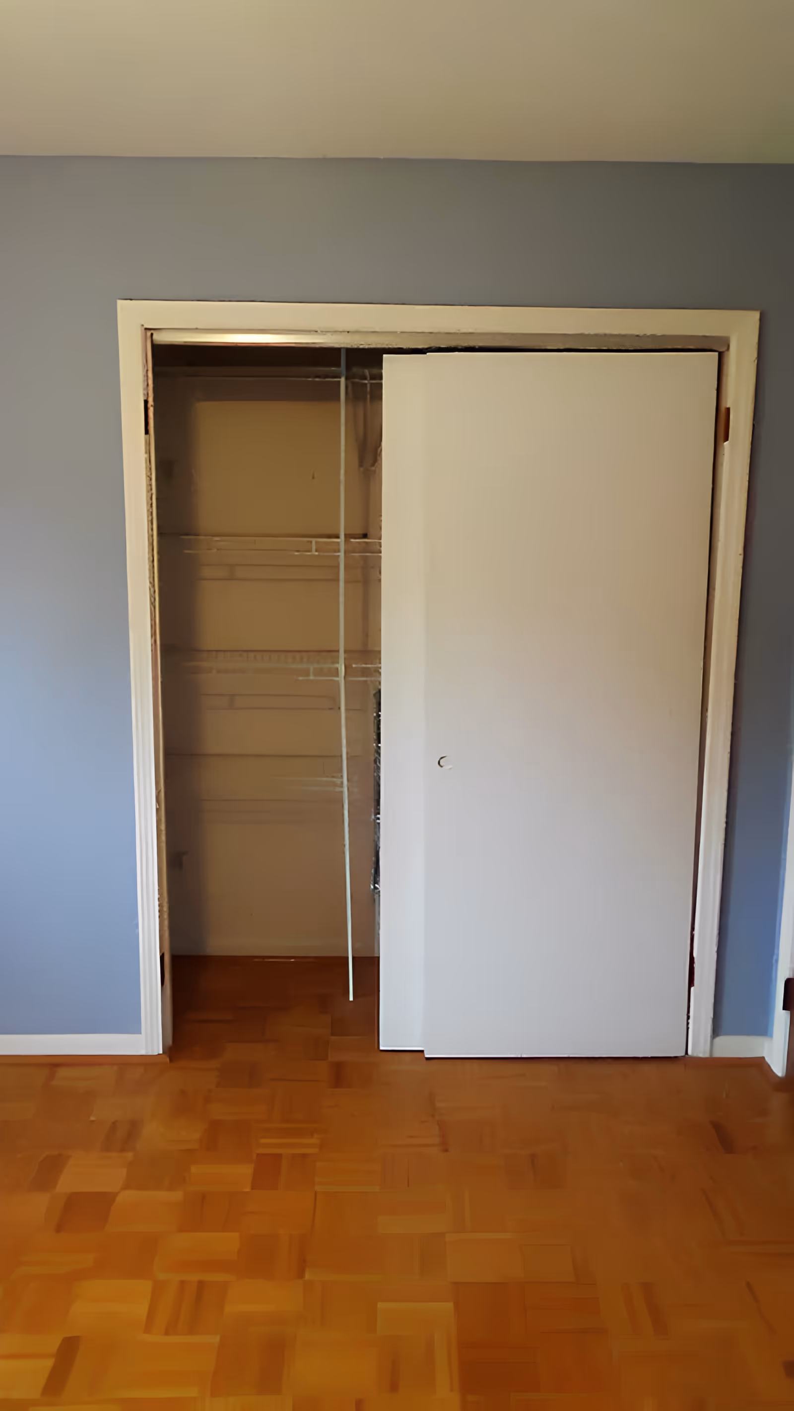 Empty room with parquet floor and a closet with a partially open white sliding door revealing wire shelving against light blue walls.