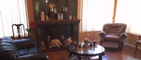 A cozy living room with a dark leather sofa on the left, a round glass coffee table in the center, and a tufted armchair near large windows with vertical blinds. The room features a wooden floor and a fireplace mantel decorated with various ornaments and small sculptures.
