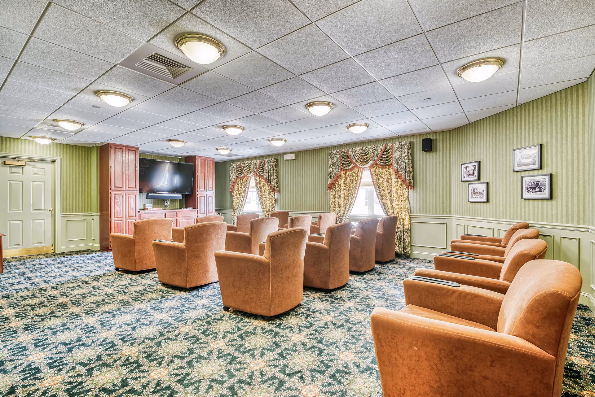 A cozy common room with multiple brown armchairs arranged in rows facing a large flat-screen TV mounted on a wooden cabinet. The room has green striped wallpaper, patterned carpet, and two windows with decorative floral curtains. Several framed pictures hang on the wall.