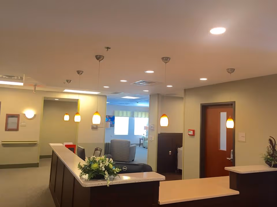 Reception area inside a senior living facility with a front desk featuring a flower arrangement, pendant lights hanging from the ceiling, and a seating area with armchairs visible in the background through an open doorway.