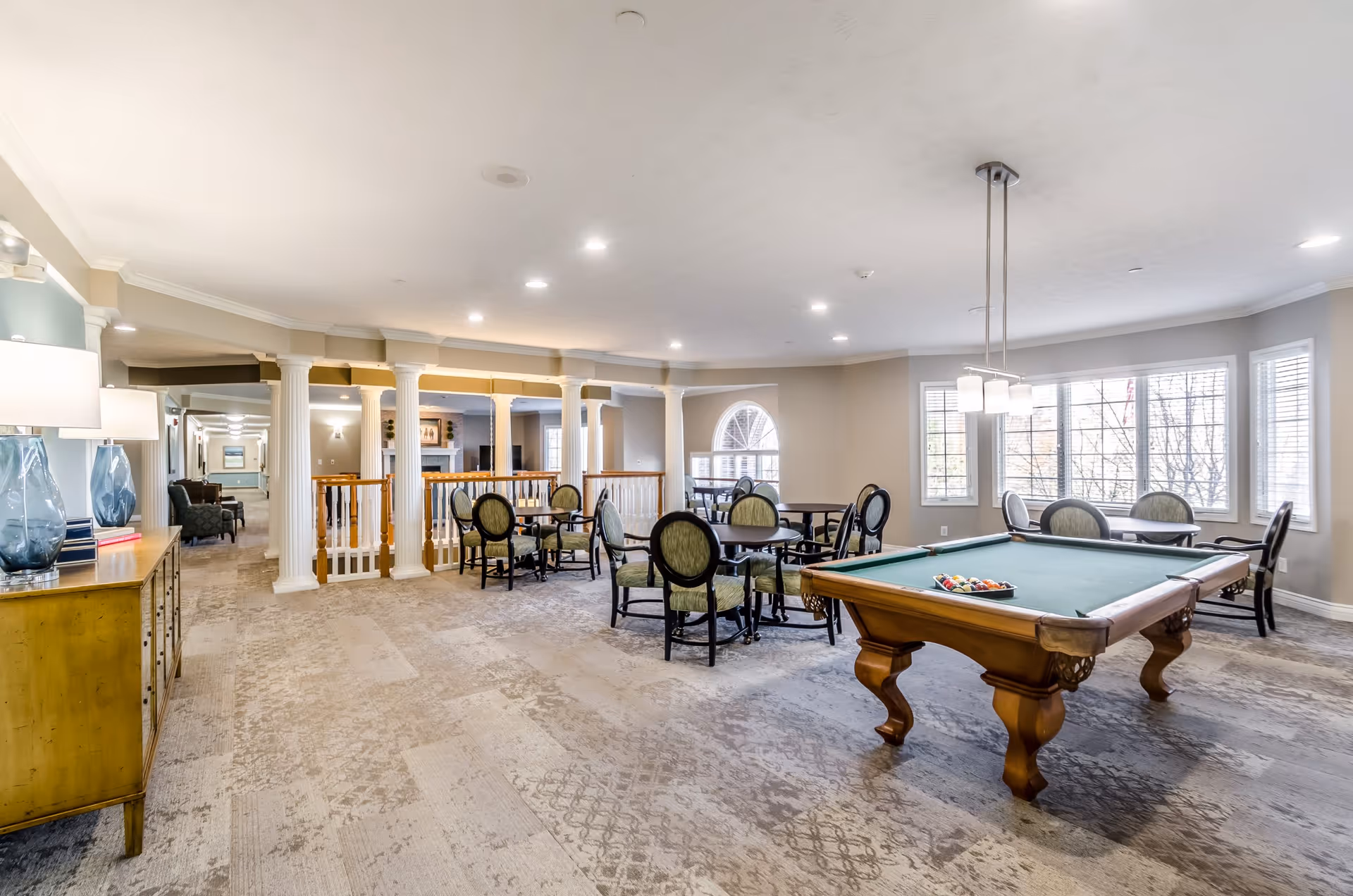 A spacious common area in an assisted living facility featuring a pool table with balls arranged for a game, several round tables with green cushioned chairs, large windows letting in natural light, and decorative columns separating the space. The room has a neutral color palette with beige walls and carpeted flooring.