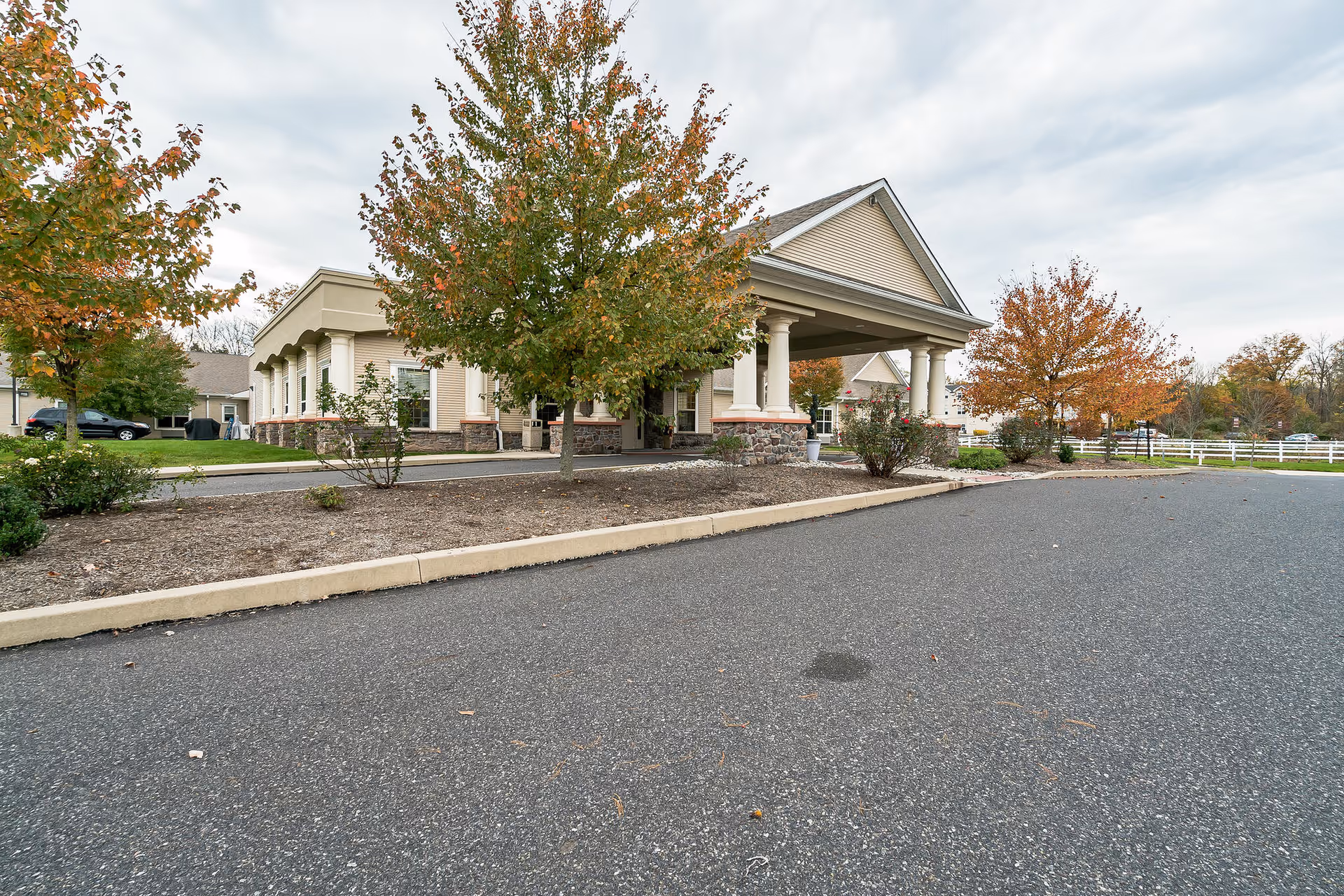 Exterior view of a senior living facility building with a covered entrance supported by columns. There are trees with autumn foliage and a paved driveway in front of the building.