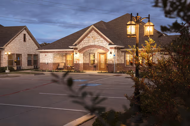 Exterior view of a single-story senior living facility building made of stone and brick with a peaked roof, illuminated by warm outdoor lights at dusk. There is a parking lot in front with marked handicap spaces and a vintage-style street lamp on the right side surrounded by bushes.