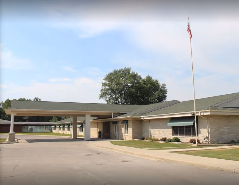 Single-story stone senior living facility with a covered entrance drive, green awnings, and a flagpole.