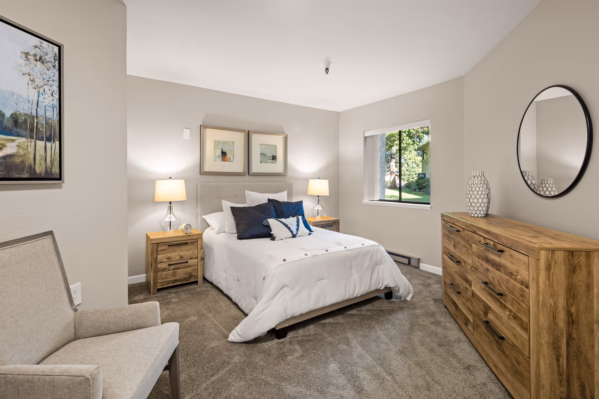 A cozy bedroom with a neatly made bed featuring white bedding and multiple pillows. There are two wooden nightstands with lamps on either side of the bed, a wooden dresser with a decorative vase and a round mirror above it, a beige upholstered chair, and a window showing greenery outside. The walls are painted light gray and the floor is carpeted.