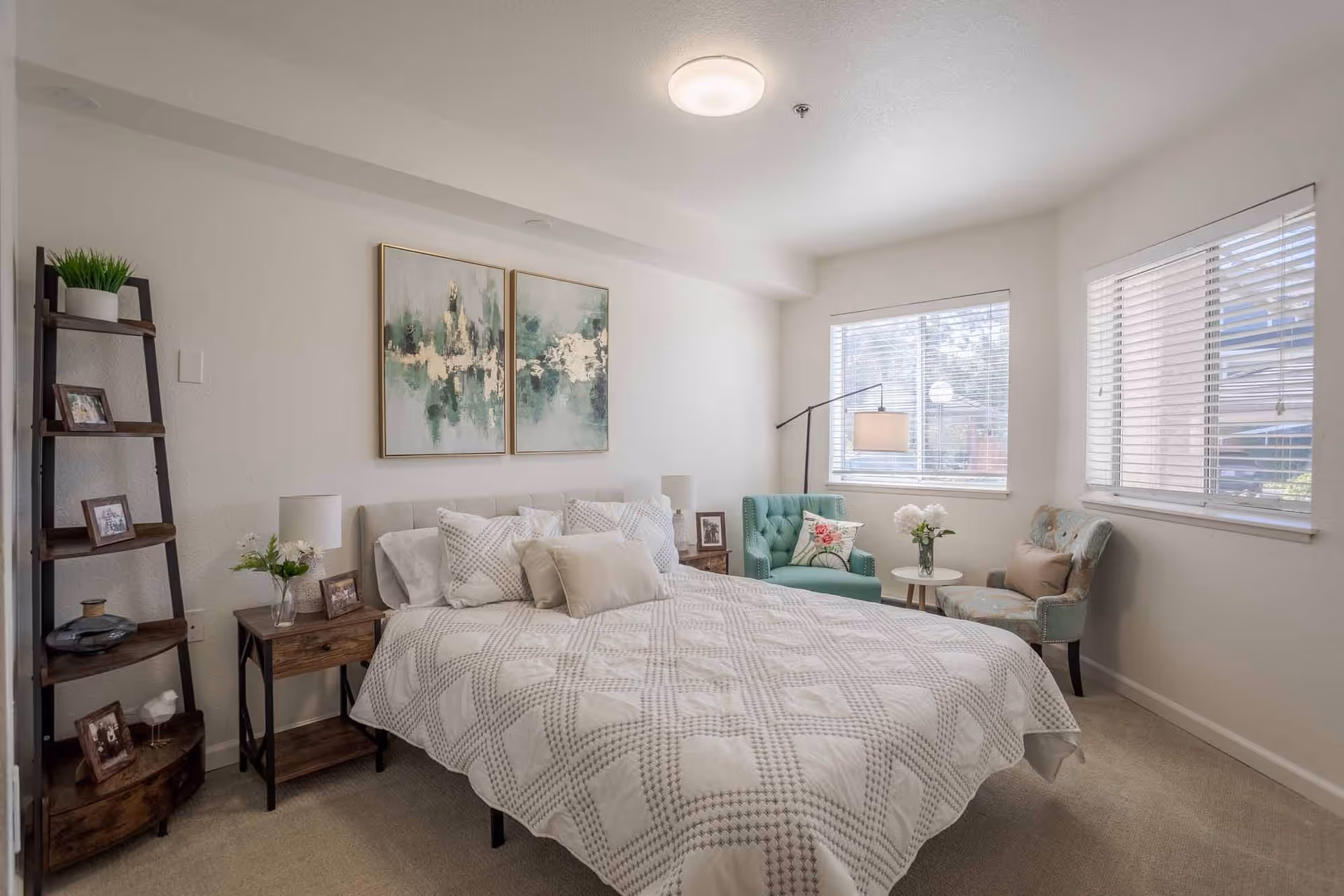 A bright and cozy bedroom with a large bed covered in a white and beige patterned quilt. The room features two windows with white blinds, a teal armchair with a floral pillow, a matching ottoman, a small round white table with a vase of flowers, and a wooden ladder shelf with framed photos and a plant. Two abstract paintings hang above the bed, and there are bedside tables with lamps and decorative items.
