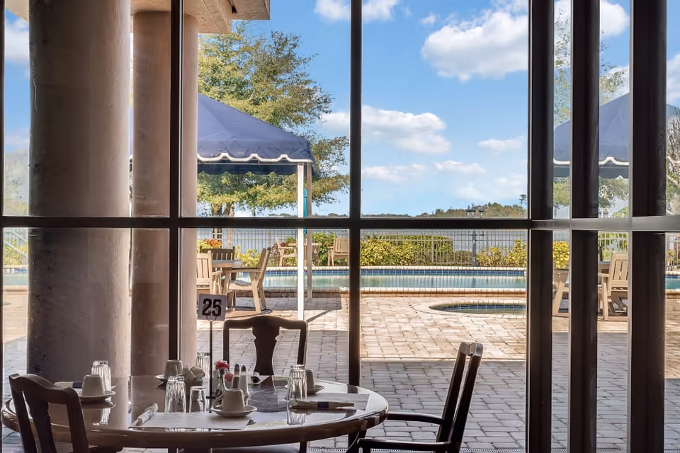 View through large glass windows of an outdoor patio area with a swimming pool, tables, chairs, and blue canopies. Inside, a round dining table is set with cups, glasses, and utensils, with a table number 25 visible.