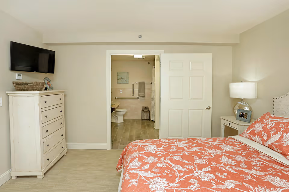 A bedroom with a coral and white floral bedspread on the bed, a white nightstand with a lamp and a framed photo, a white dresser with a basket and a photo frame on top, and a wall-mounted TV. The room has beige walls and carpeted floor. An open door reveals an accessible bathroom with a toilet, grab bars, and a shower curtain.