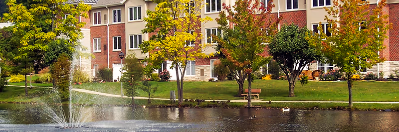 Exterior of a multi-story senior living building with a pond and fountain, trees, and a bench along the shore.