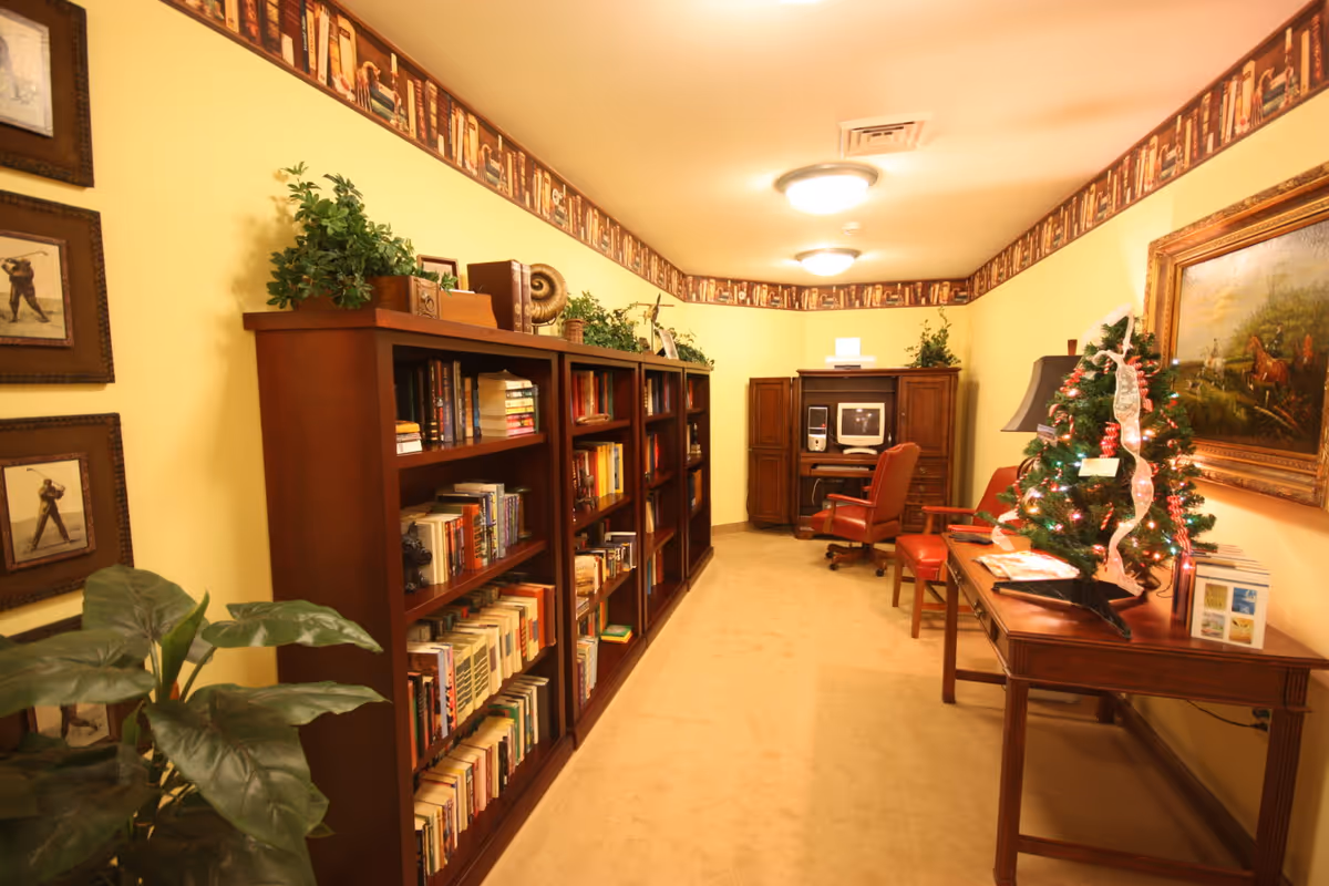 Long cozy reading room with wooden bookshelves lining the left wall, a desk with a small decorated Christmas tree on the right, and seating near a computer at the far end.