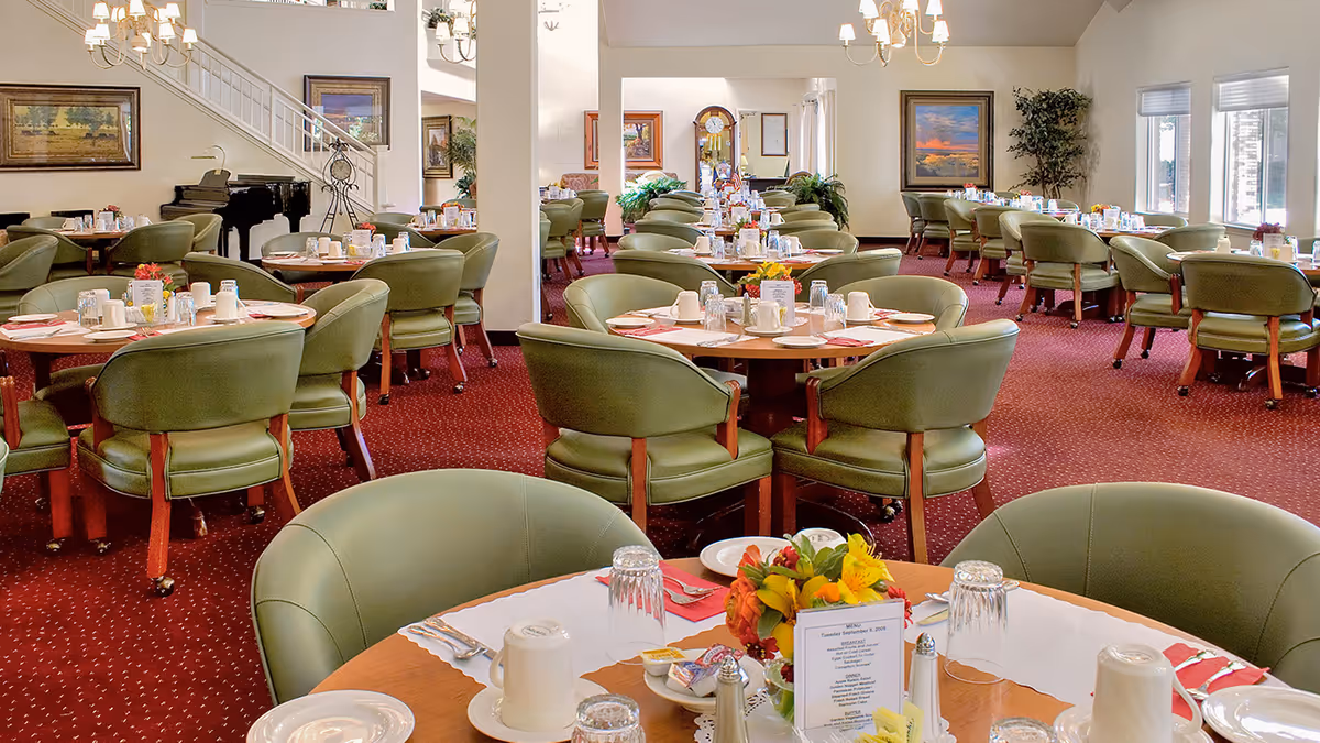 Spacious dining room with round tables set for meals, green upholstered chairs, and chandeliers.