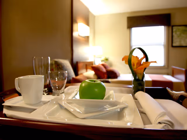 A close-up view of a tray with a white coffee cup, a glass, a green apple on a white plate, and a small vase with orange flowers in a cozy room with a bed and pillows in the background.