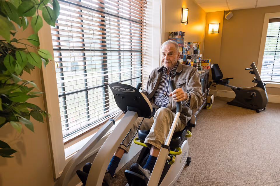 An elderly man using a recumbent exercise bike in a well-lit room with large windows covered by blinds. The room has beige walls, carpeted floor, a green plant near the window, and another exercise bike in the background along with a cabinet stacked with board games.