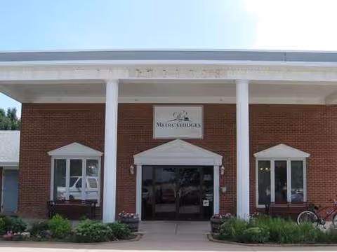 Brick Medicalodge of Kinsley facility with white columns, glass entrance, benches, and planters.
